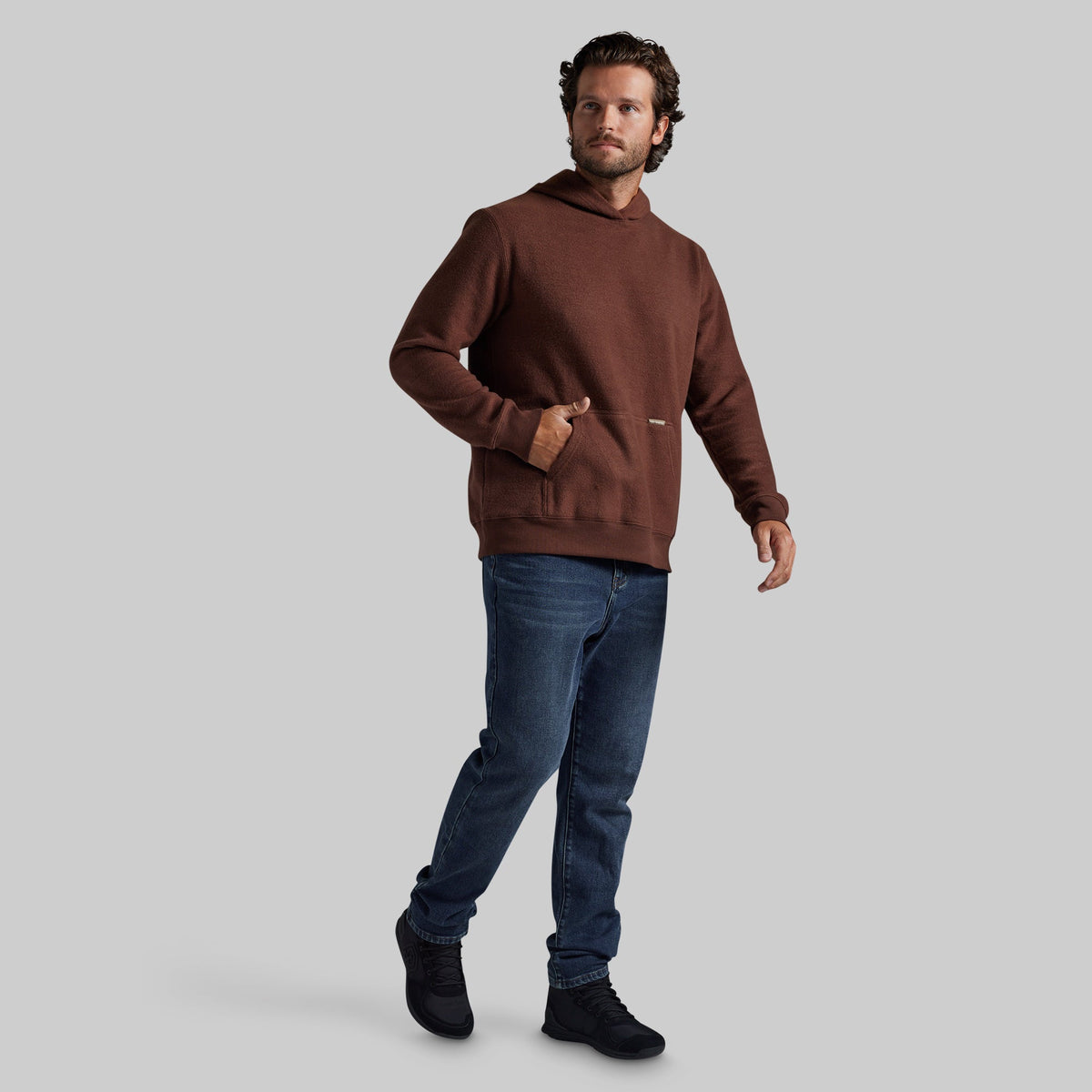 A man modeling a brown hooded sweatshirt with blue jeans and black shoes, standing against a solid gray studio background.