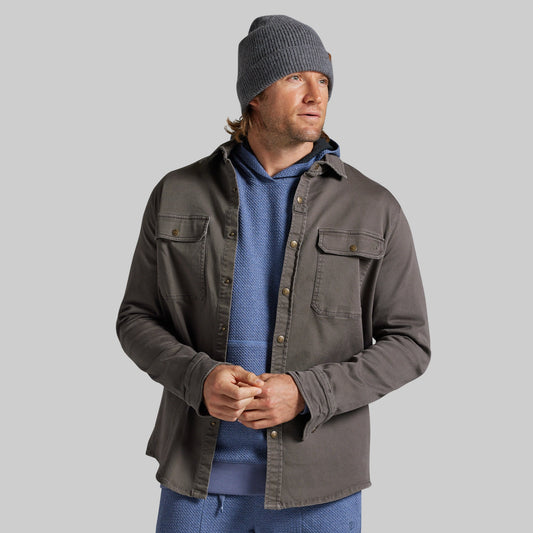 A man wearing a dark gray button-up overshirt over a blue knit hoodie and matching blue pants, paired with a gray beanie, photographed against a solid gray studio background.