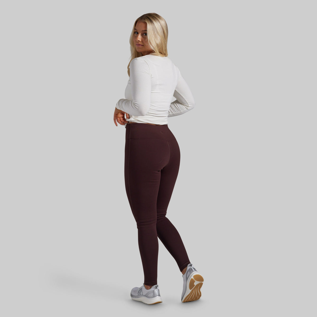 A woman is shown from the back wearing a white long-sleeve athletic top and dark brown leggings, paired with gray and tan sneakers, against a solid light gray background.