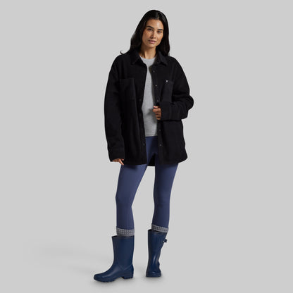 A woman modeling a black sherpa fleece jacket over a gray top, blue leggings, and blue rain boots against a solid gray studio background.