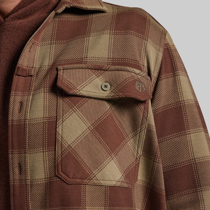 Close-up of a brown and beige plaid flannel shirt jacket with a chest pocket and snap tab, layered over a brown hoodie against a light gray background.