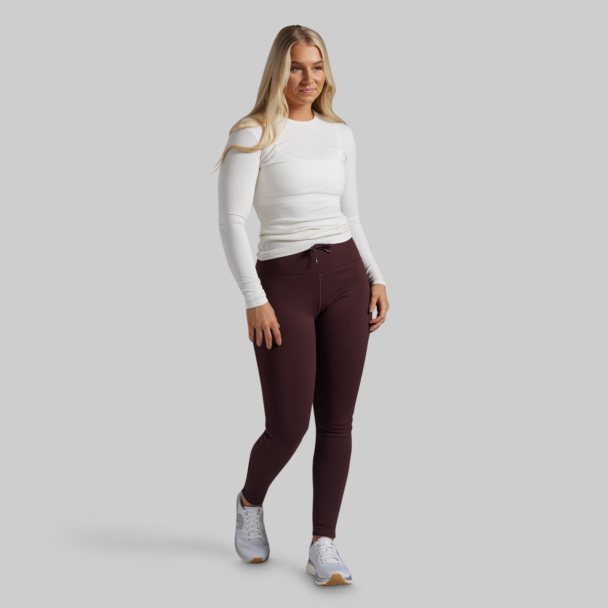 A woman modeling a white long-sleeve top and dark burgundy leggings with white sneakers, standing against a solid light gray background.