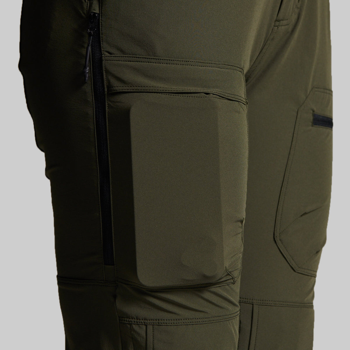 Close-up of olive green technical outdoor pants showing a large front flap pocket, side zippered pocket, and angled cargo pocket on the thigh.