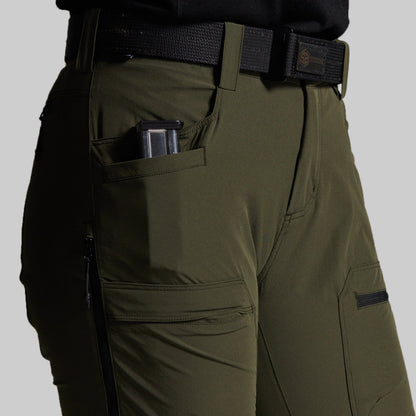 Close-up of olive green outdoor pants worn with a black belt, showing side pockets, a zippered pocket, and a handgun magazine in the side pocket against a light gray background.