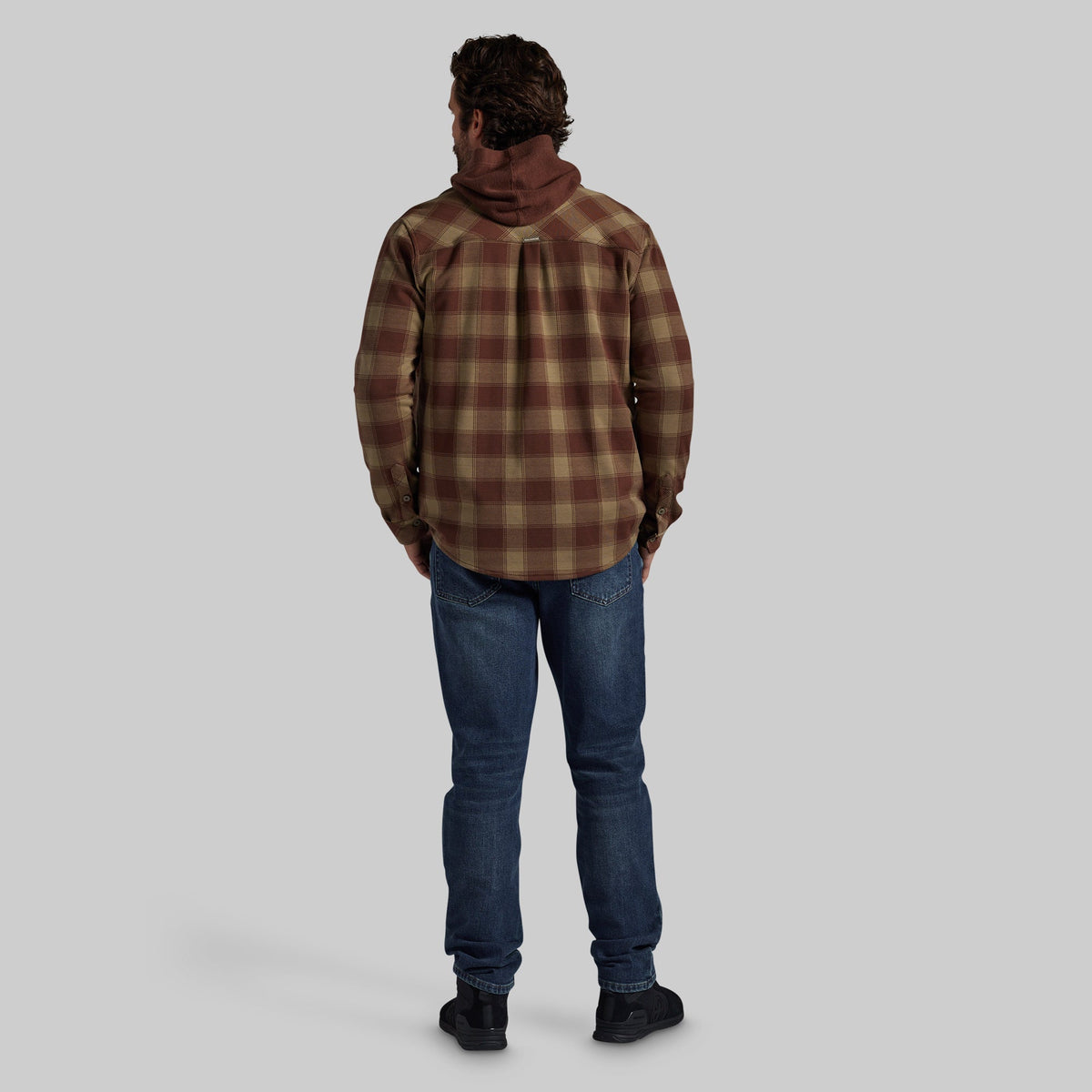 A man viewed from behind wearing a brown and tan plaid hooded flannel shirt, blue jeans, and black shoes, standing against a solid gray studio background.