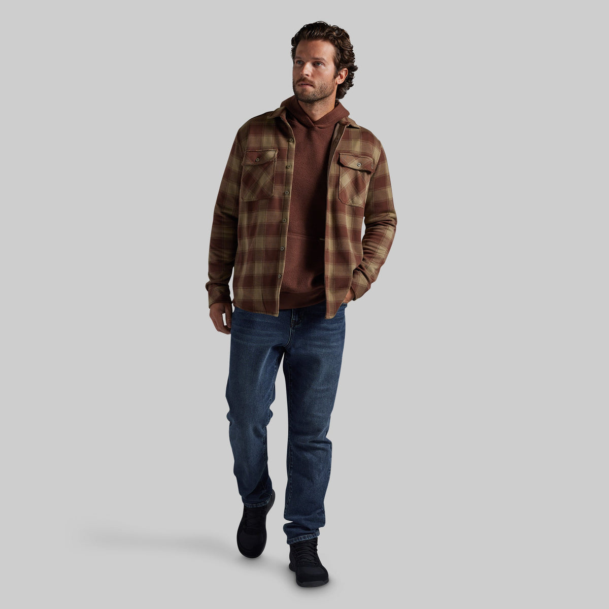 A man modeling a brown and tan plaid flannel shirt jacket over a brown hoodie, paired with blue jeans and black boots, against a solid gray background.