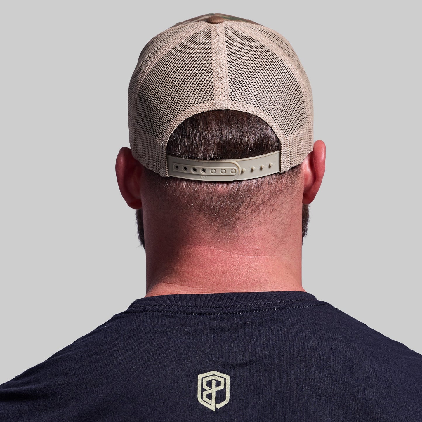 Person wearing a beige mesh cap and navy shirt with a logo on a gray background