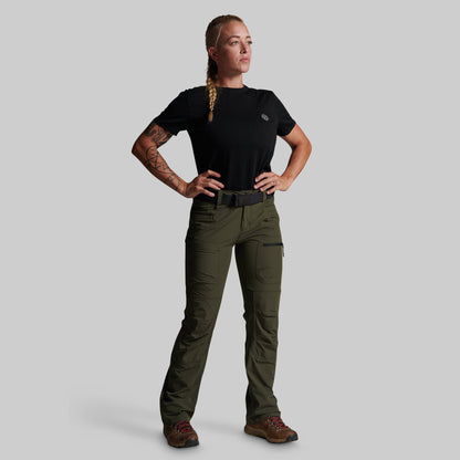 A woman modeling a pair of olive green outdoor cargo pants with a black t-shirt and brown hiking boots, standing against a solid gray background.