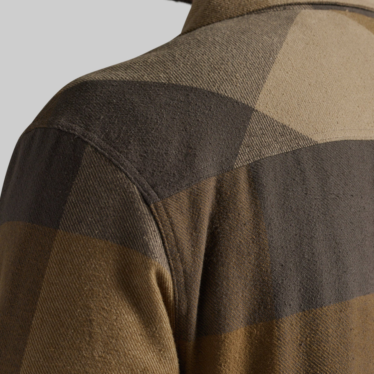 Close-up of a plaid flannel jacket fabric in earthy brown, tan, and dark charcoal tones, showing the textured woven material and shoulder seam.