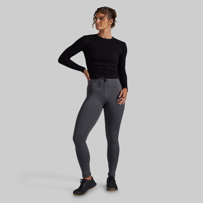 A woman modeling dark gray athletic leggings and a black long-sleeve top against a solid gray studio background.