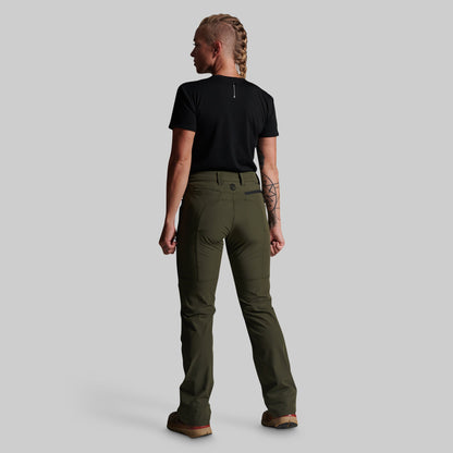 A woman shown from the back wearing olive green outdoor pants, a black t-shirt, and brown boots against a solid gray studio background.