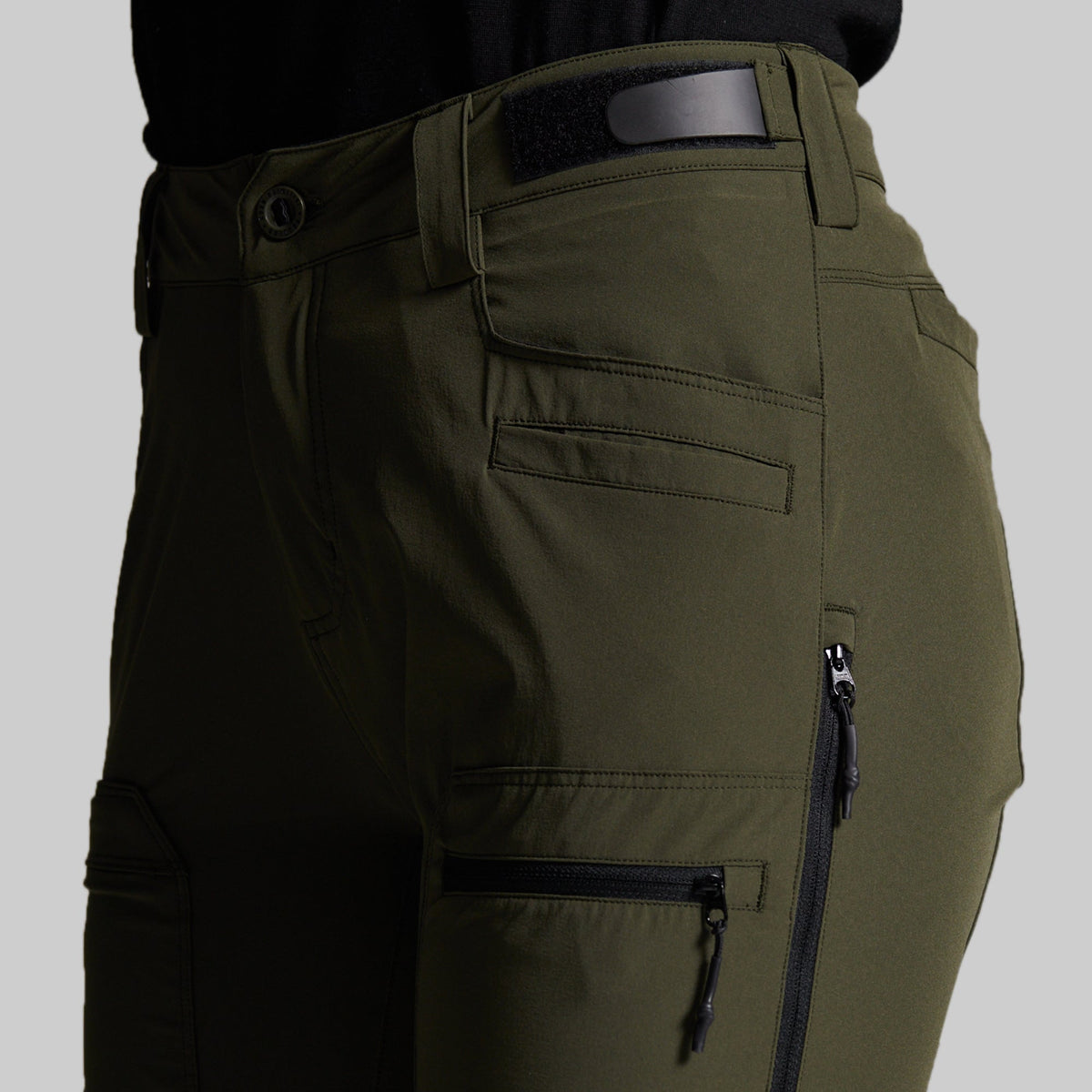 Close-up of olive green cargo pants with multiple zippered pockets and belt loops, shown on a model against a light gray background.