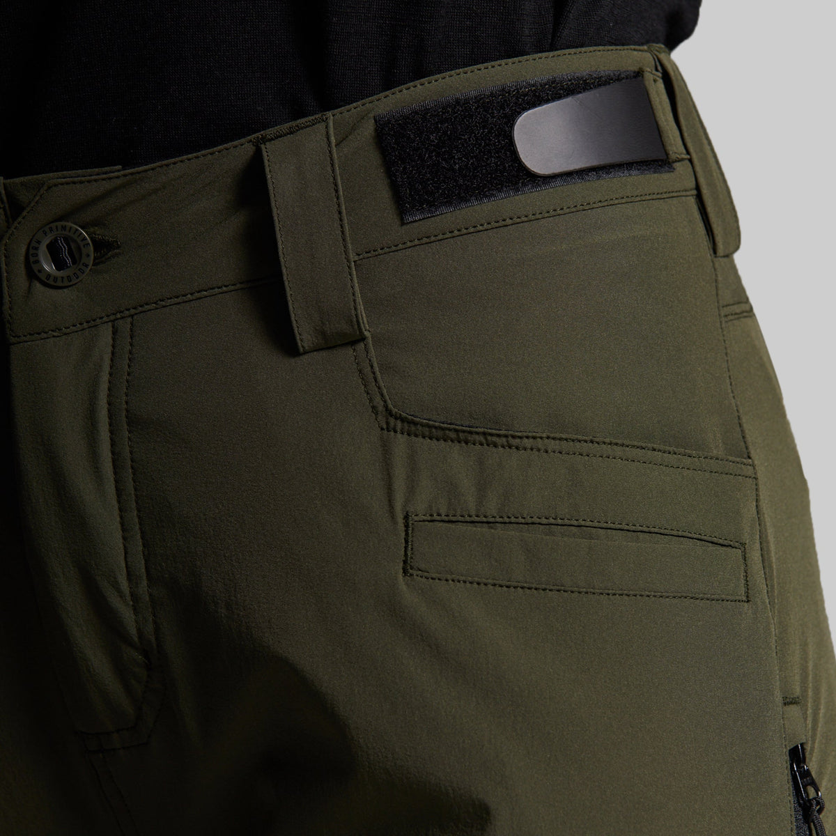 Close-up of olive green outdoor pants showing the waistband, belt loop with a black insert, button closure, and front pocket stitching.