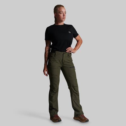 A woman modeling olive green cargo pants with a black t-shirt and brown hiking boots, standing against a solid gray background.