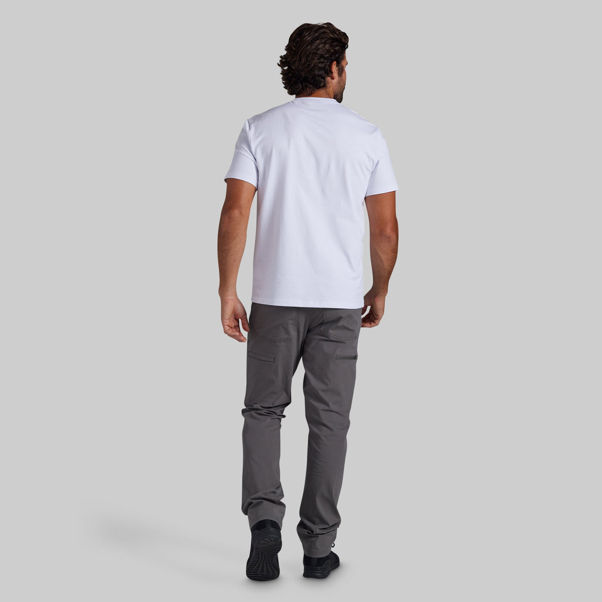 A man viewed from behind wearing a white short-sleeve t-shirt and dark gray pants, standing against a solid light gray background.