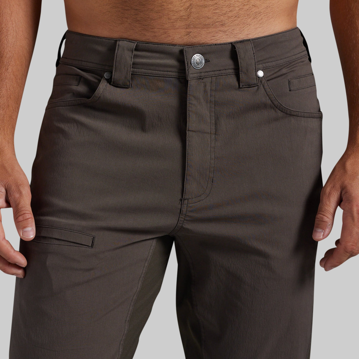 Close-up of a person wearing dark brown cargo-style pants, showing the waistband, front pockets, and thigh cargo pocket against a light gray background.