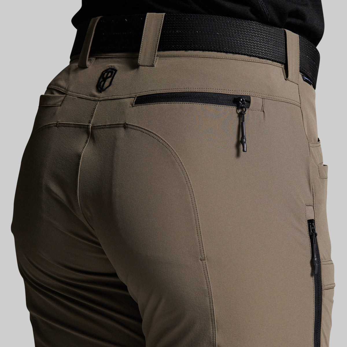 Close-up of the back waist and pockets of storm grey outdoor pants worn with a black belt, showing zippered pockets and a small circular logo emblem.