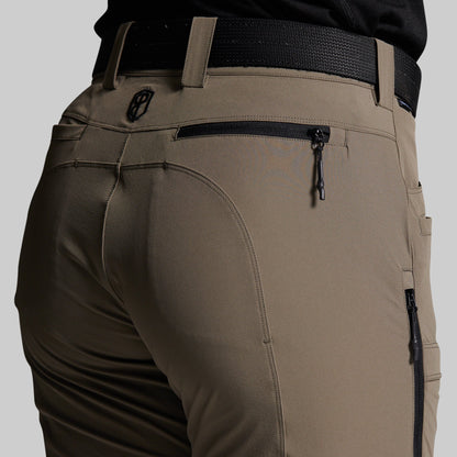 Close-up of the back waist and pockets of storm grey outdoor pants worn with a black belt, showing zippered pockets and a small circular logo emblem.