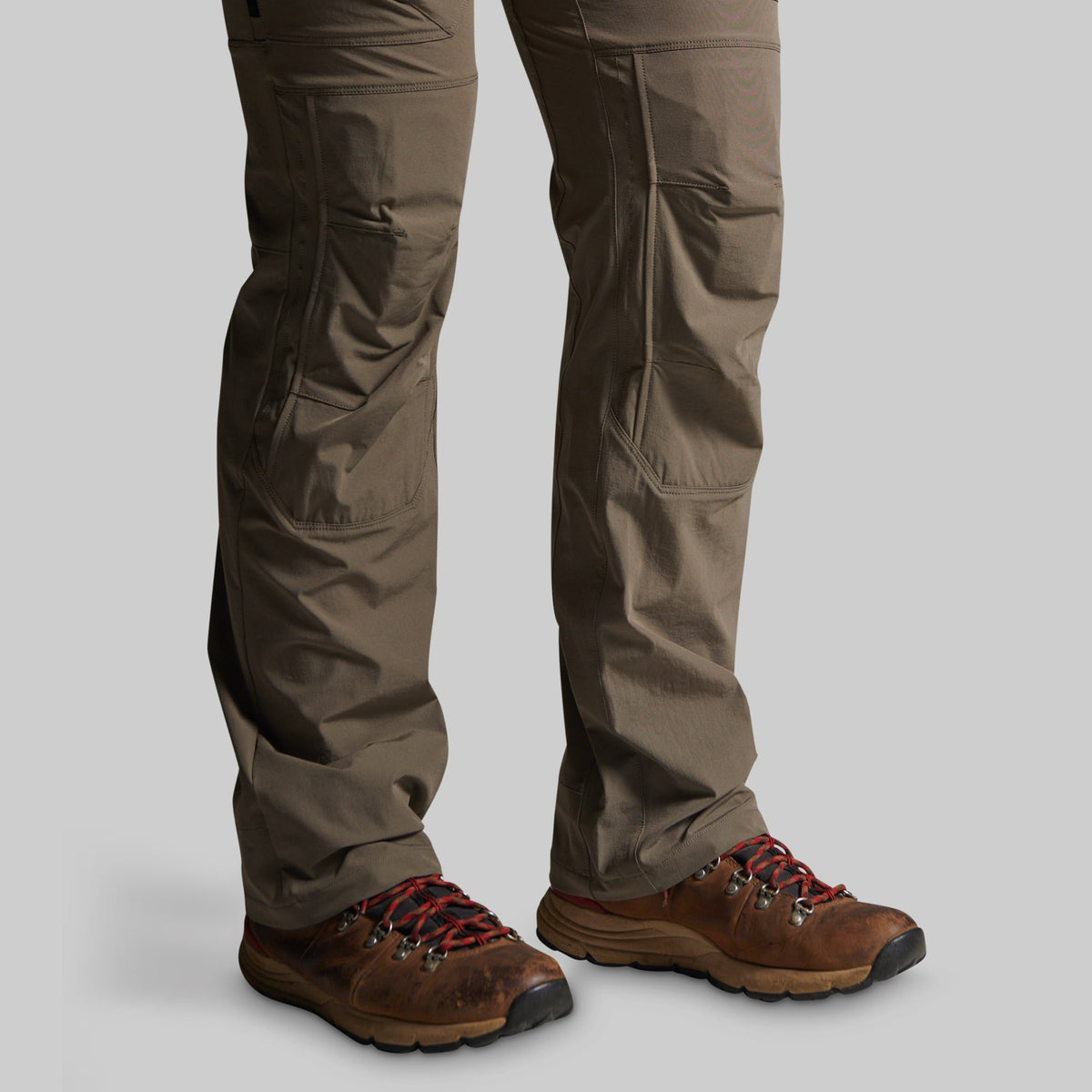A close-up of a person's legs wearing brown hiking boots and storm grey cargo pants against a solid gray background.
