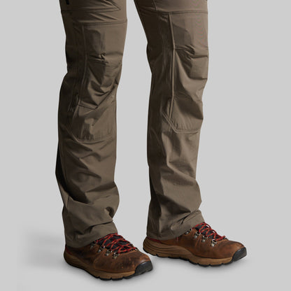 A close-up of a person's legs wearing brown hiking boots and storm grey cargo pants against a solid gray background.