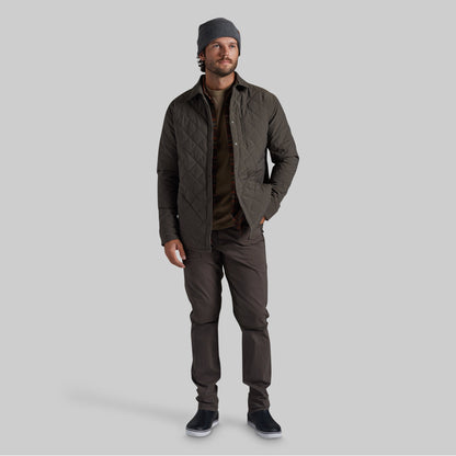 A man wearing a dark olive green quilted jacket, brown shirt, dark brown pants, a gray beanie, and black slip-on shoes, standing against a solid gray studio background.