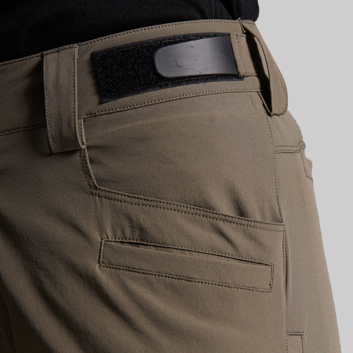 Close-up of a person wearing storm grey outdoor pants with a black belt and a black waistband patch, shown against a light gray background.