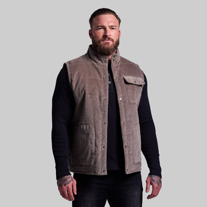 Man wearing a brown vest over a black long-sleeve shirt on a gray background
