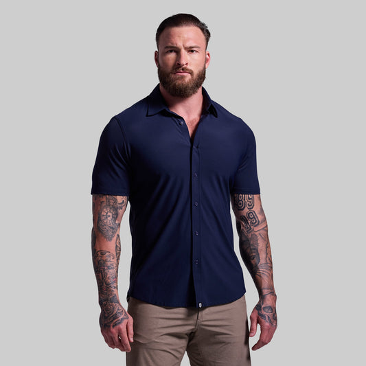 A man with tattoos wearing a navy blue short sleeve button-down shirt paired with light-colored pants.