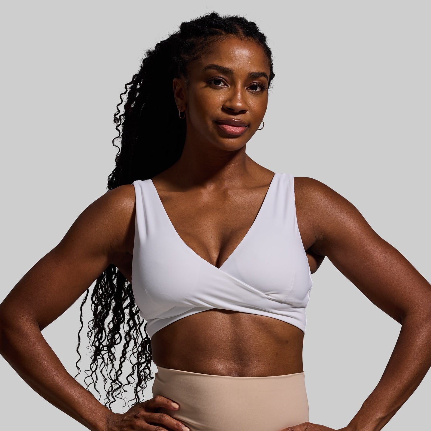 Woman wearing a white sports bra and beige pants against a gray background