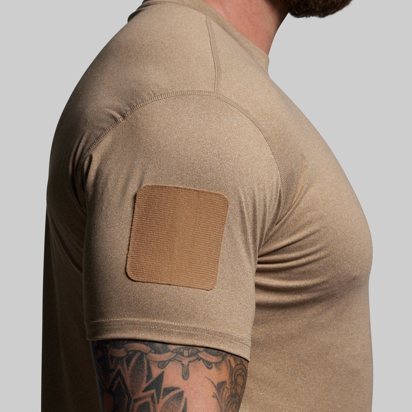 Person wearing a brown tactical shirt with a patch on a gray background