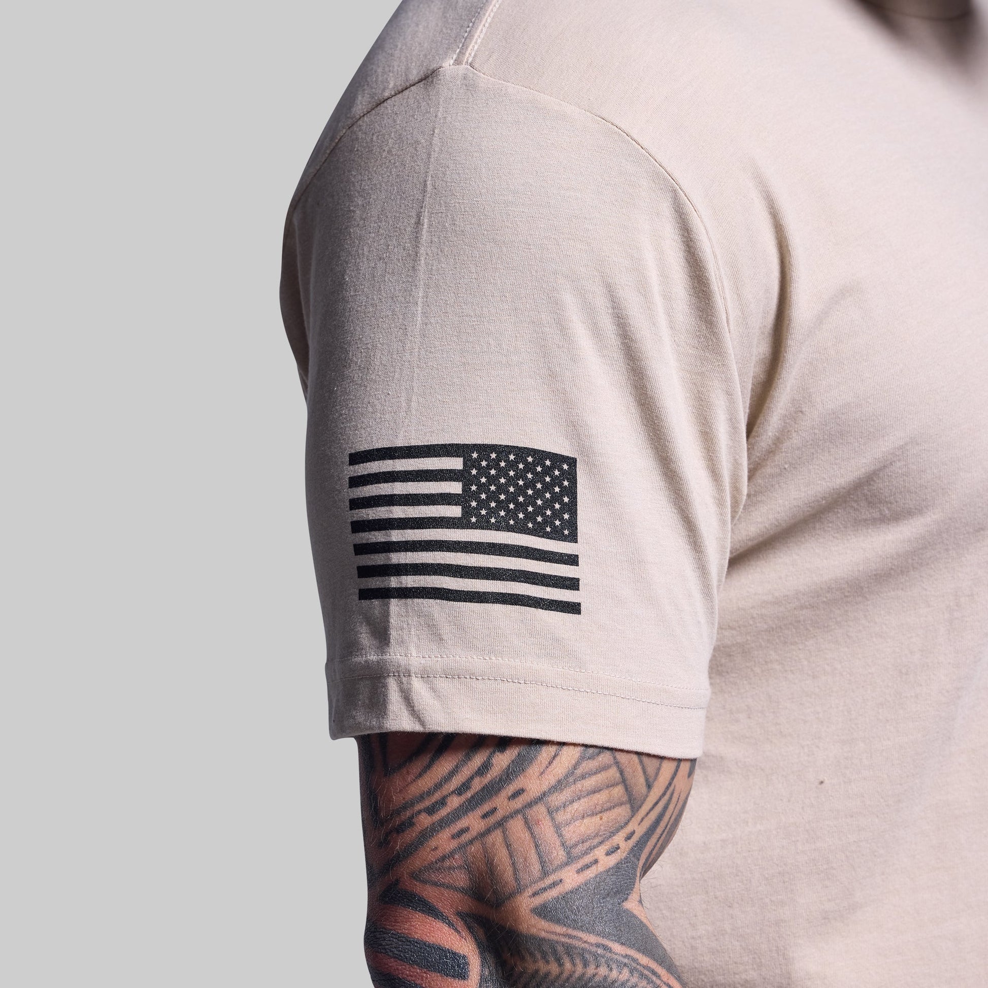 Beige t-shirt with American flag patch on sleeve against a gray background