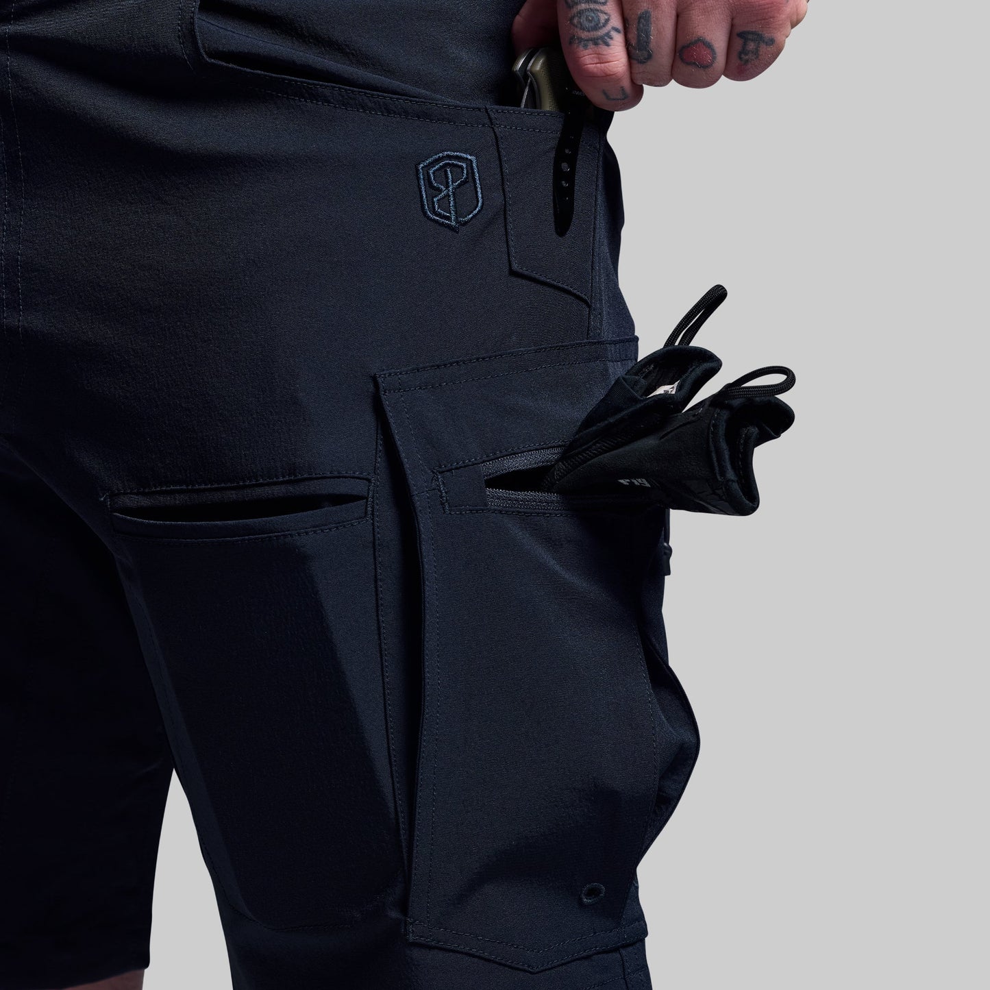Person wearing dark blue shorts with a visible brand logo with multiple objects in pockets, on a light gray background