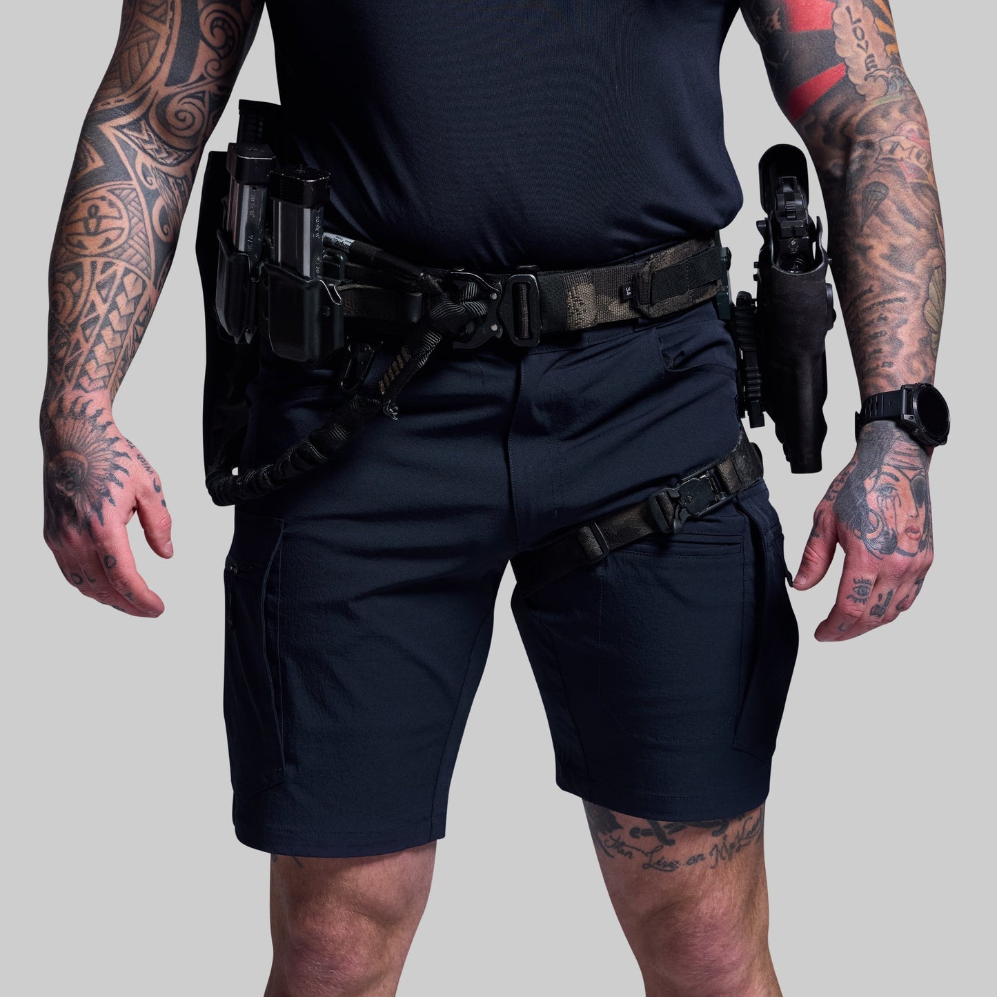 Person wearing blue tactical shorts and gear with tattoos on arms and hands on a gray background