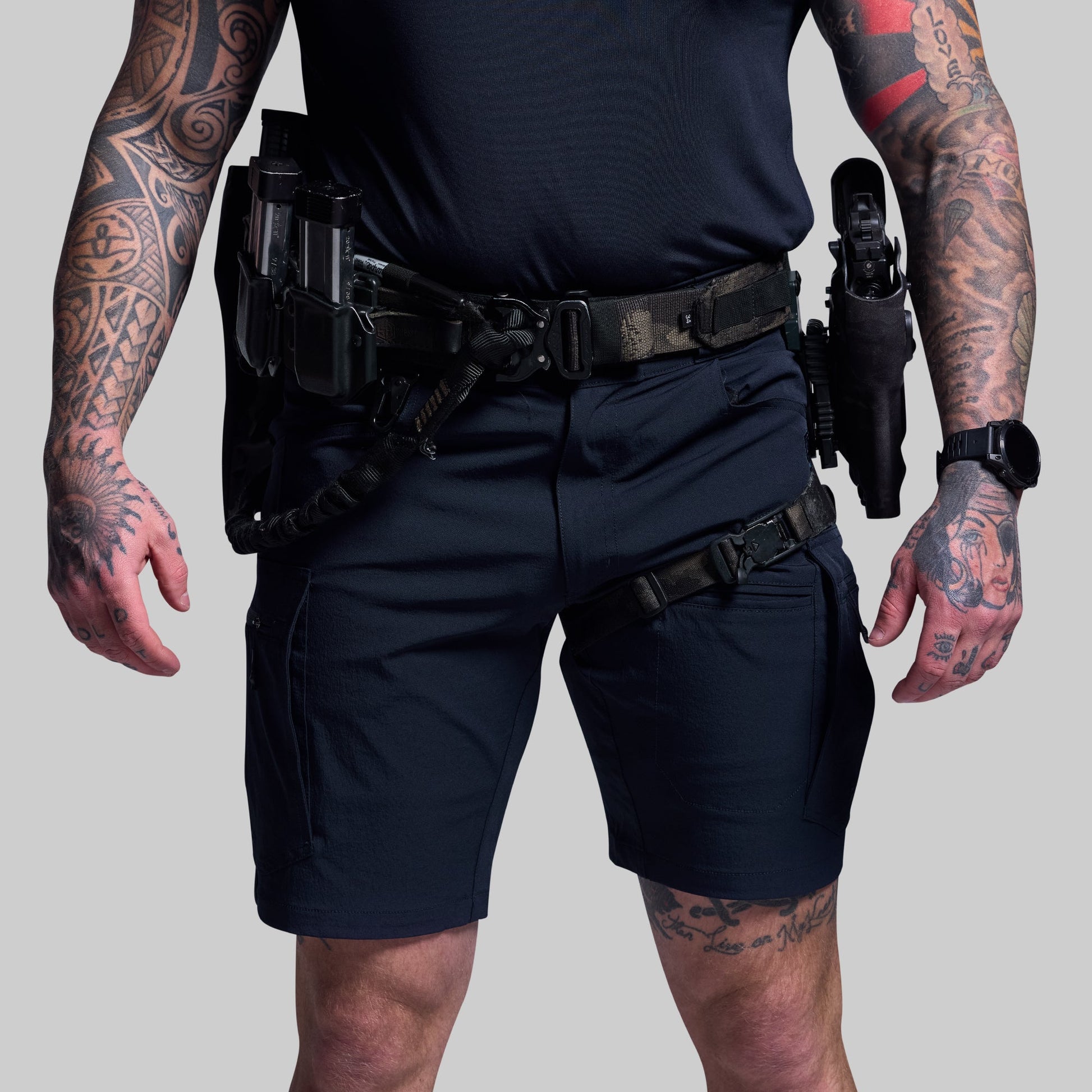 Person wearing blue tactical shorts and gear with tattoos on arms and hands on a gray background