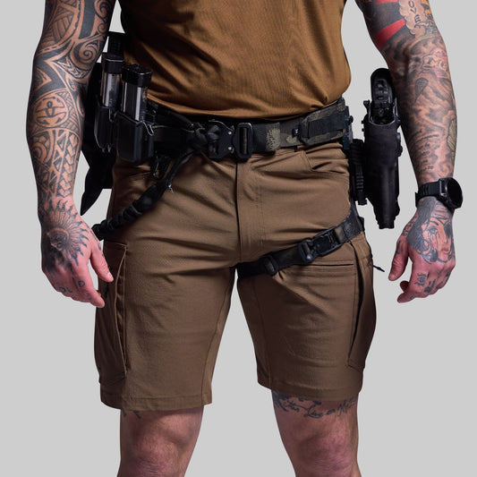 Person wearing brown tactical shorts with tattoos on arms and legs, holding holstered weapons.