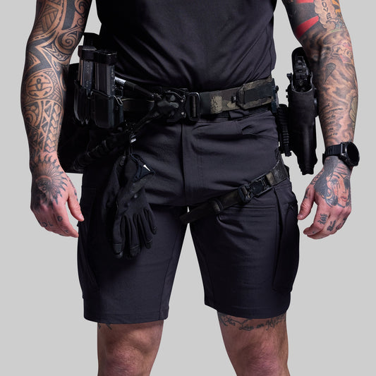 Person wearing black tactical shorts and gear with tattoos on arms and legs against a gray background