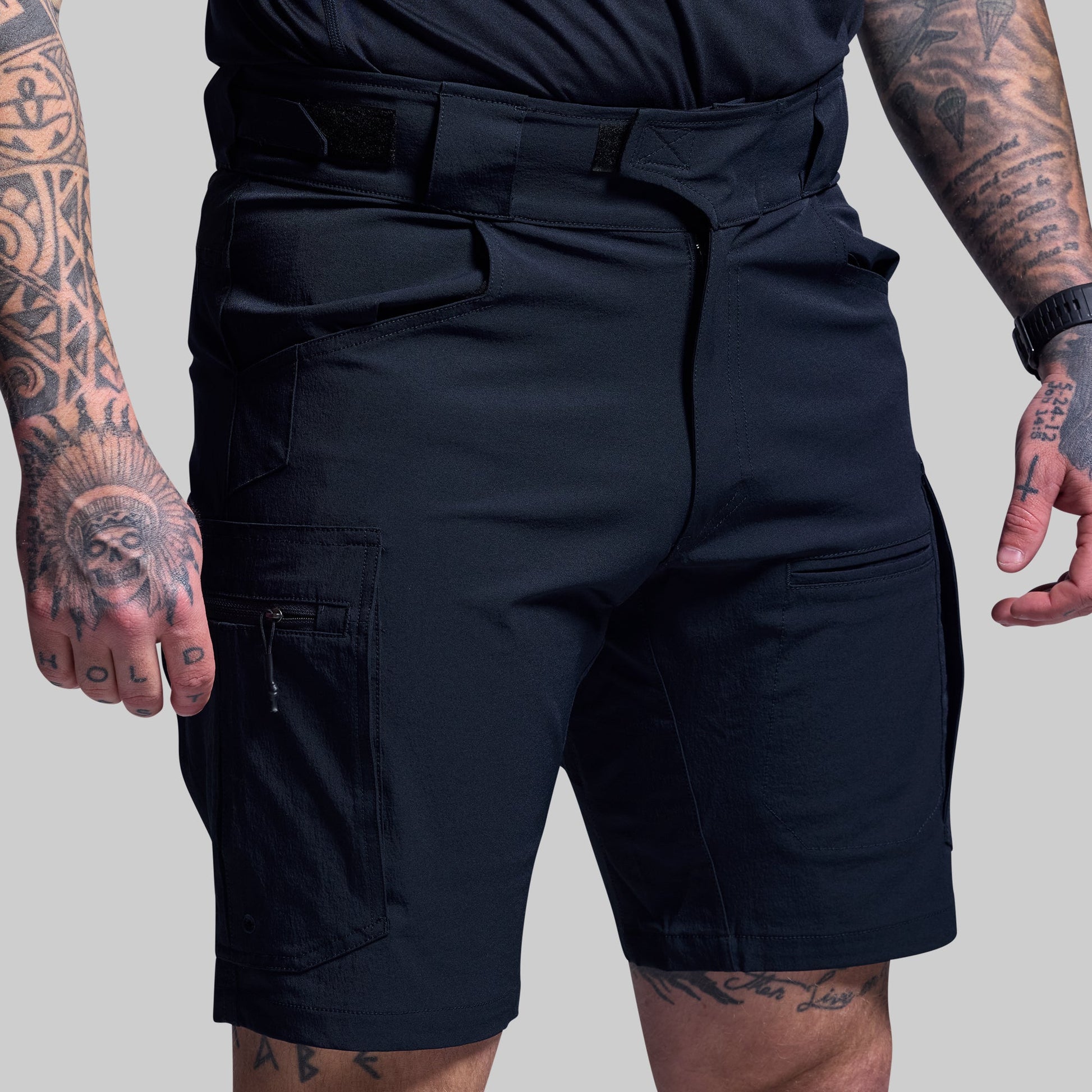 Person wearing blue tactical shorts with tattoos on arms and legs against a gray background