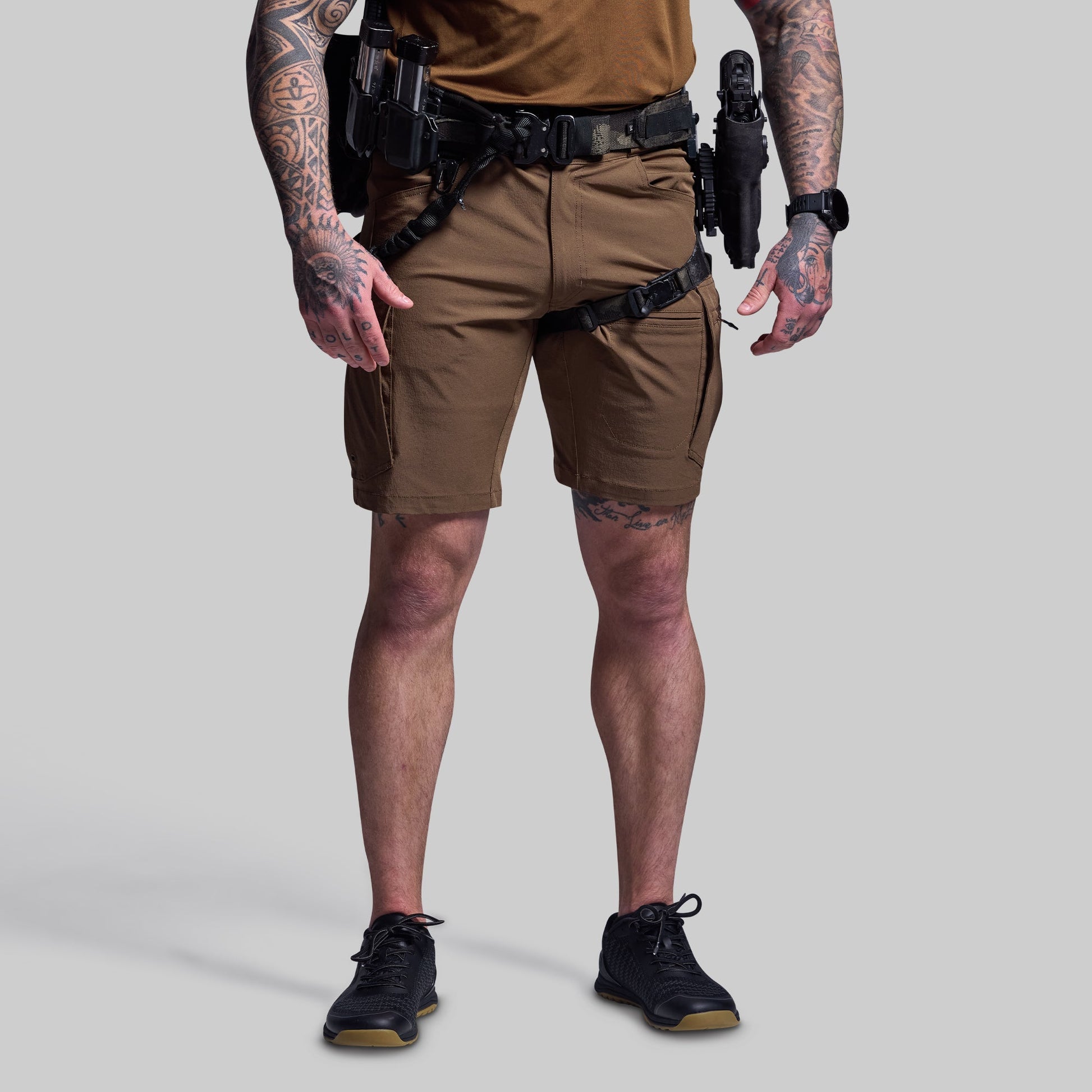 Person wearing brown tactical shorts with visible tattoos on arms and legs, on a plain background