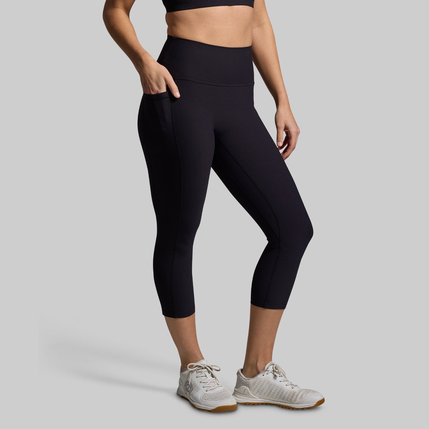 A woman wearing black capri leggings with pockets, standing upright with one hand in a pocket and wearing white sneakers.