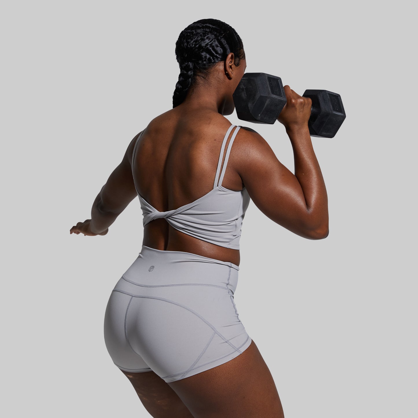 Person lifting a dumbbell in gray sports bra and shorts set on a plain background