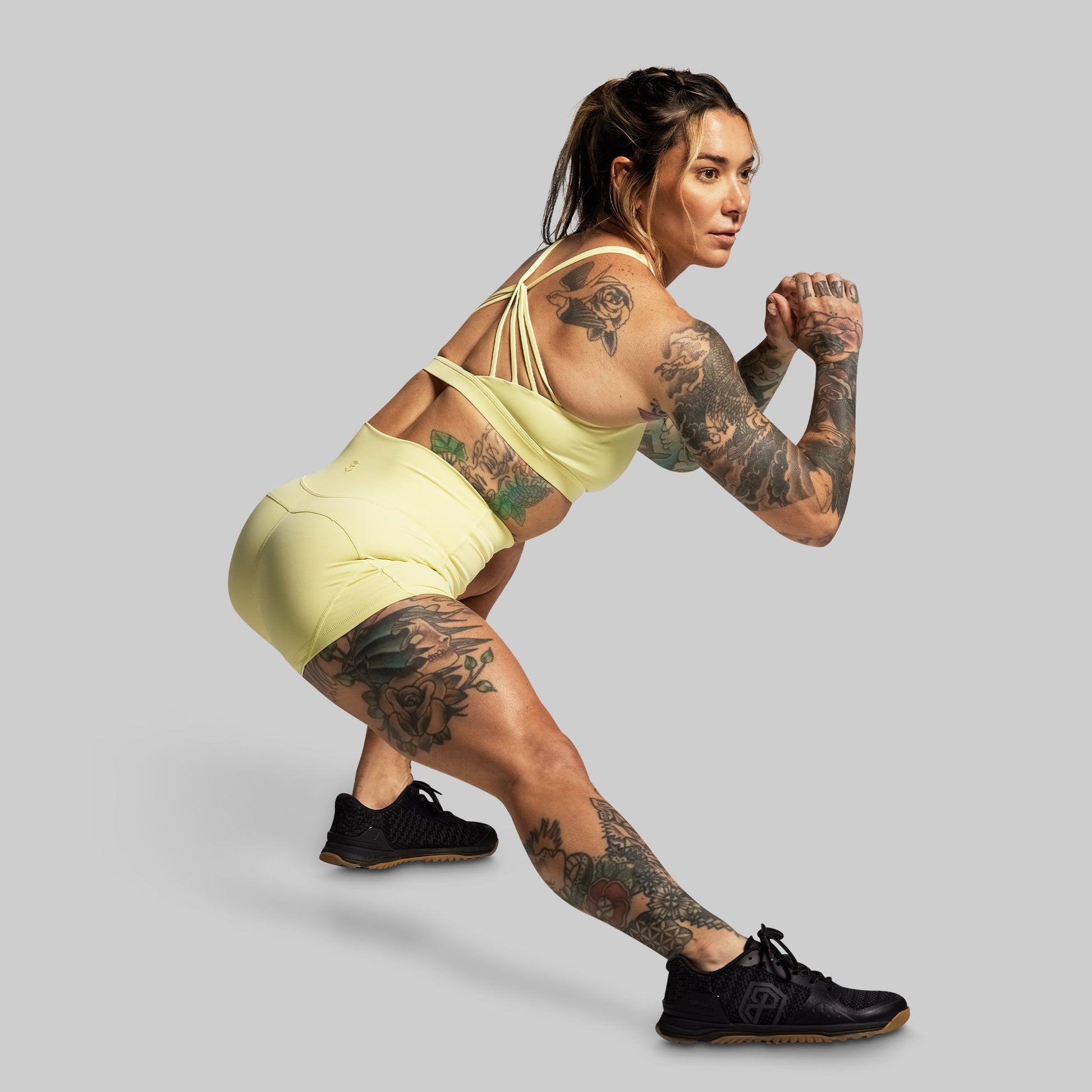 Woman in a yellow sports outfit with tattoos performing a lunge on a gray background