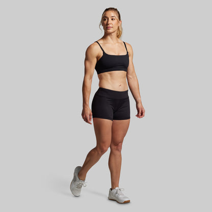 A female model wearing a black sports bra and matching black booty shorts, standing against a solid background.