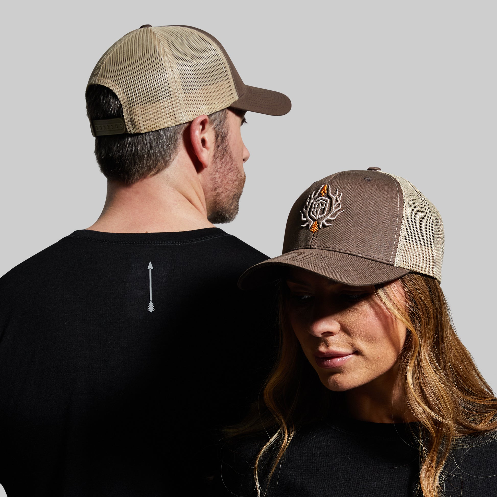 Two people wearing brown and beige trucker hats with a logo on a gray background