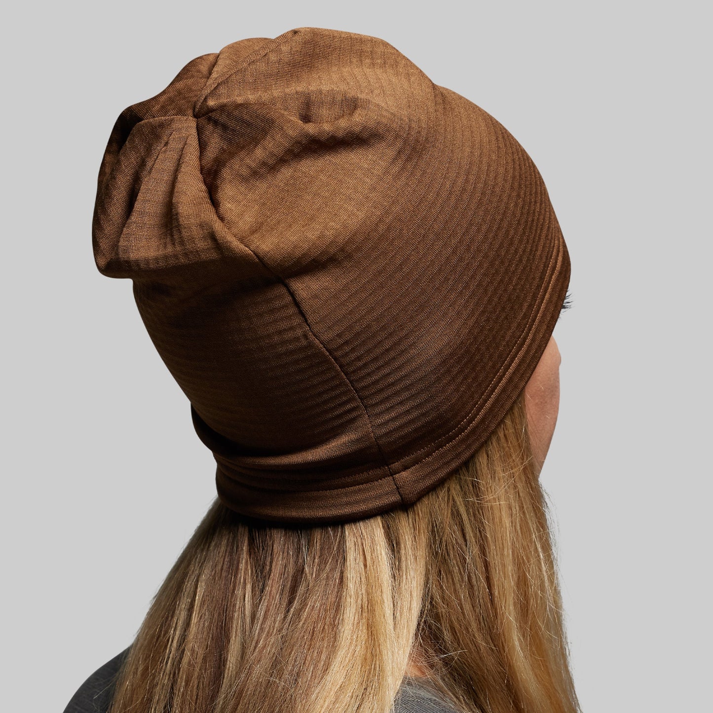 Brown ribbed knit beanie worn by a person with blonde hair on a gray background