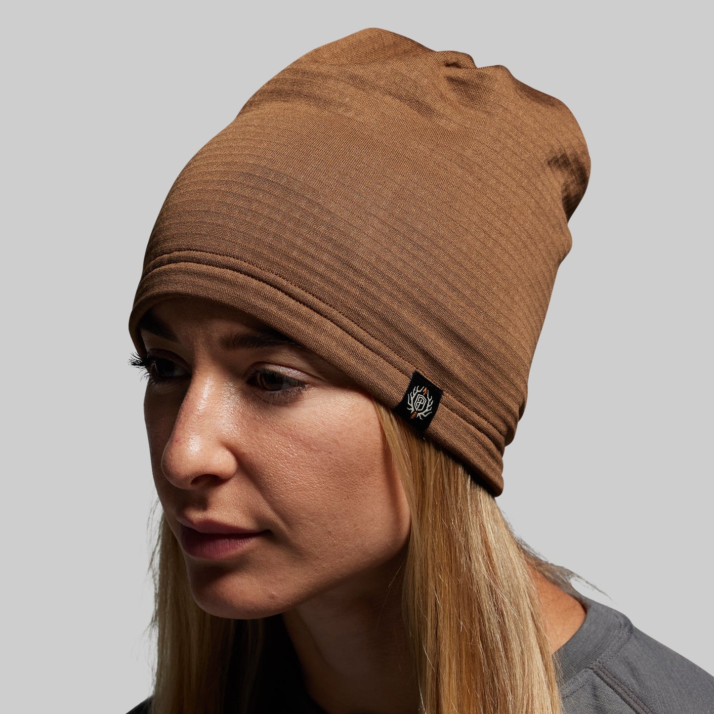 Woman wearing a brown beanie with a brand logo on a gray background