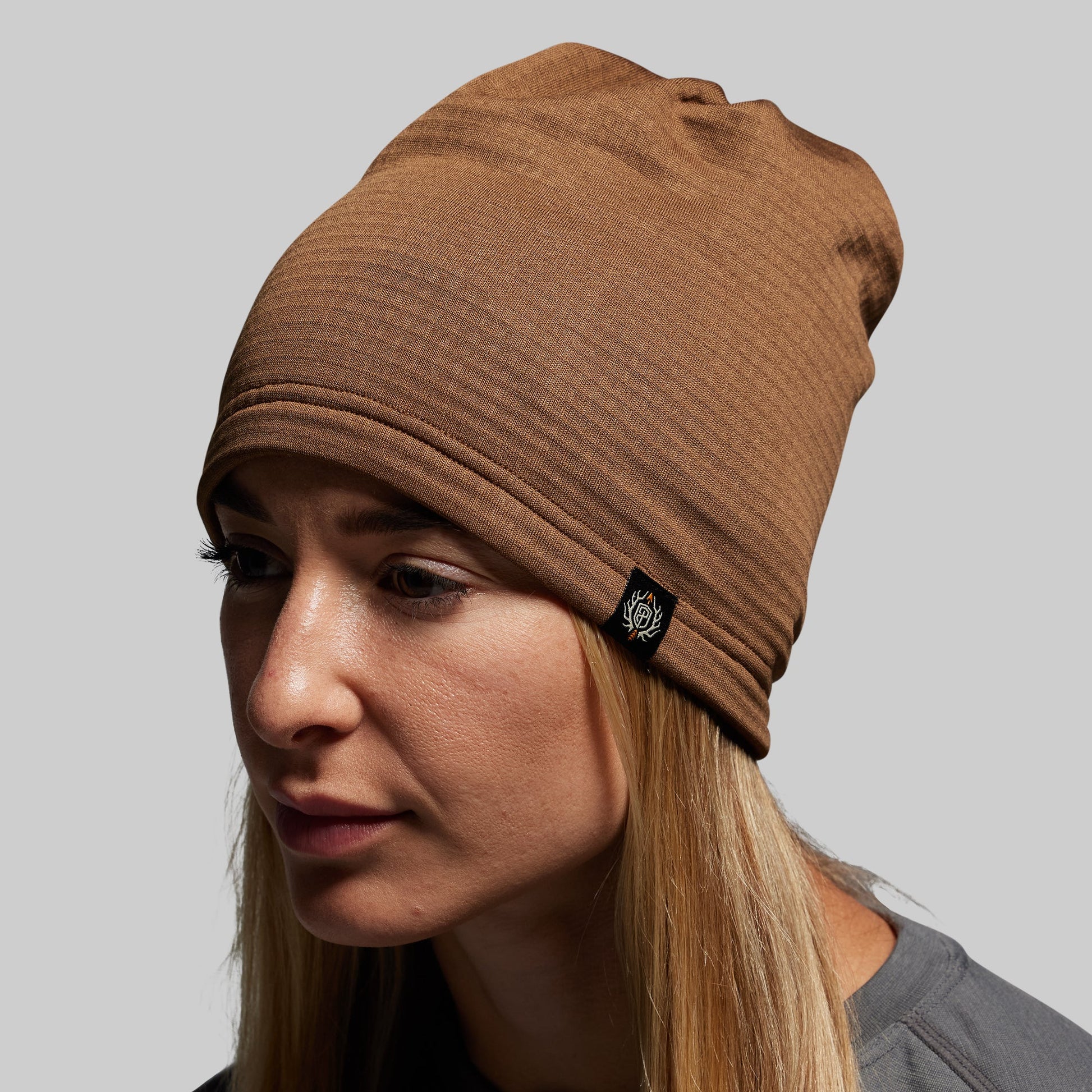 Woman wearing a brown beanie with a brand logo on a gray background