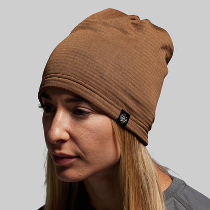Woman wearing a brown beanie with a brand logo on a gray background