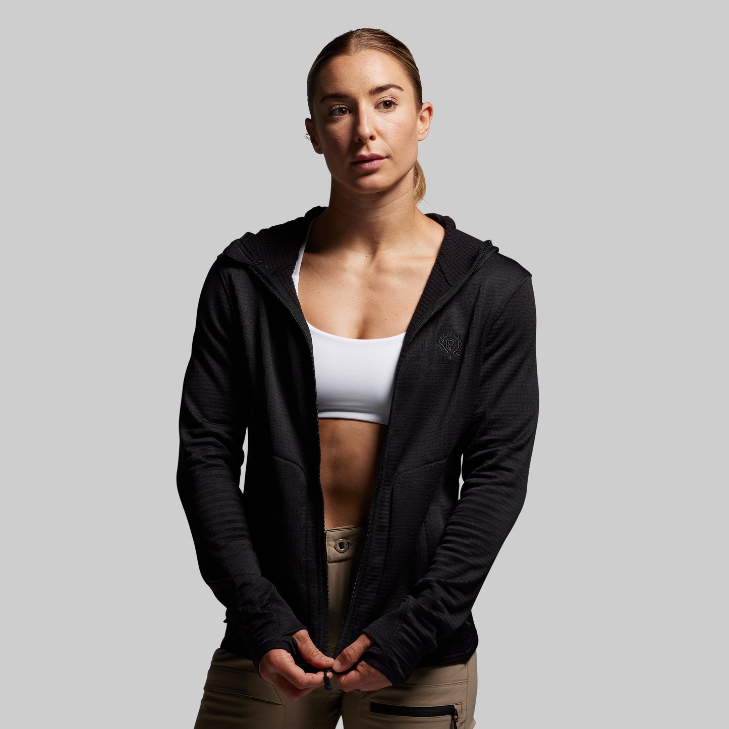 Woman wearing a black hoodie over a white sports bra and beige pants on a gray background