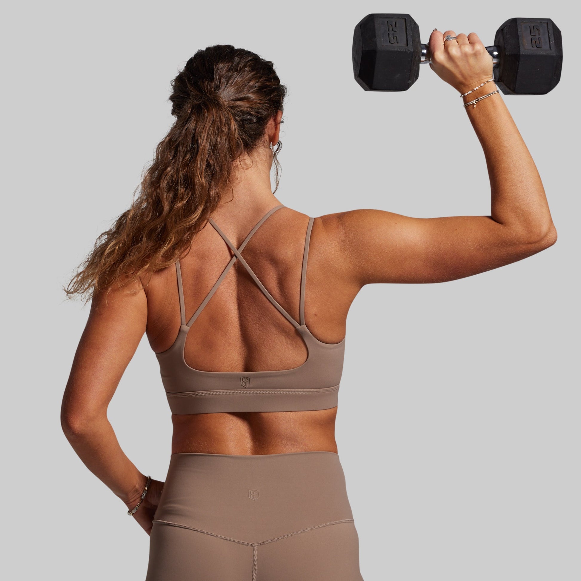 Woman in a brown, strappy sports bra lifting a dumbbell against a plain background