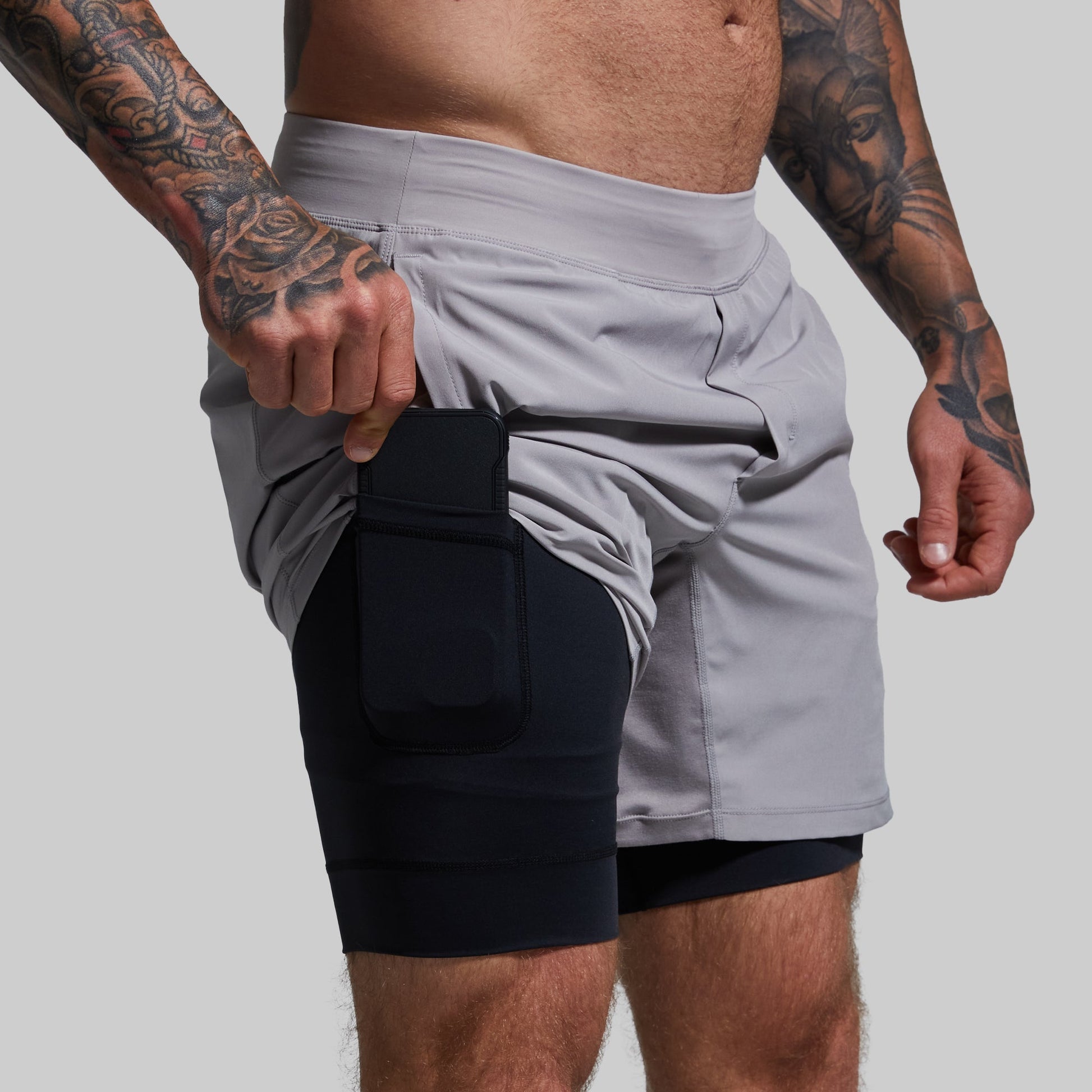 A person wearing light gray versatile shorts with a black compression liner and a phone held in the pocket.
